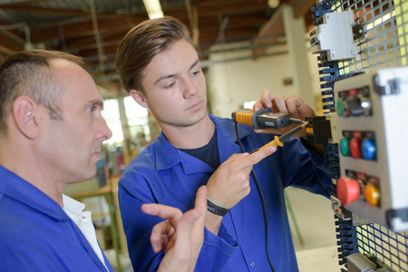 Nahaufnahme: Ausbilder beaufsichtigt Elektroniker in Ausbildung bei Arbeit an Schaltwand.